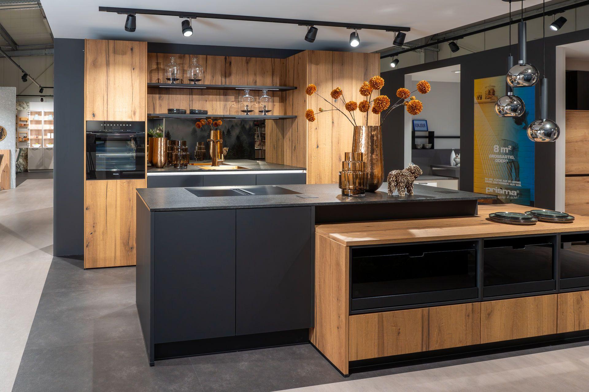 Küche in Eiche und Anthrazit mit Kochinsel, dunkler Arbeitsplatte, offenem Sideboard und Blick in die Ausstellung.