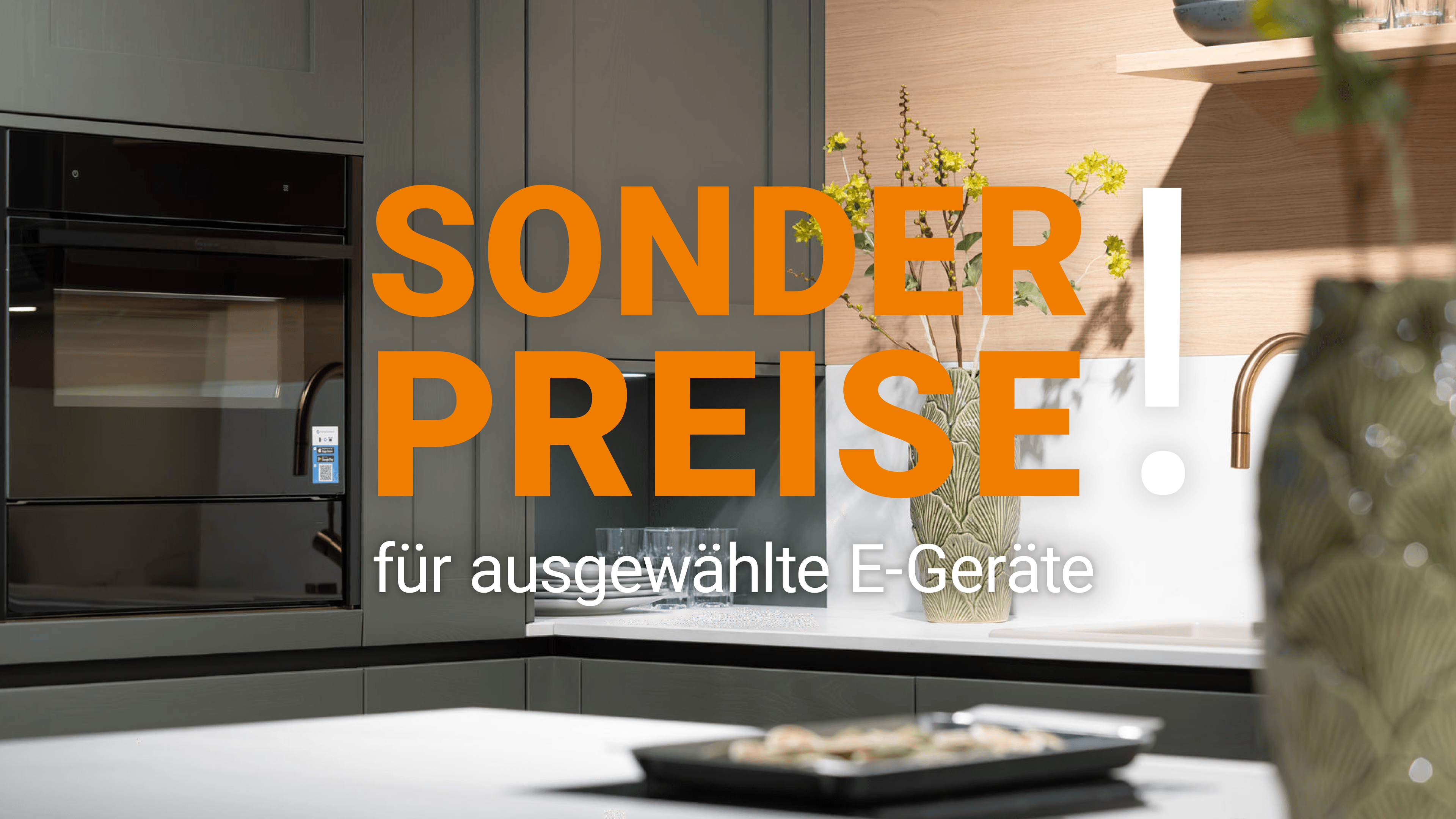 Dunkelgrüne Küche mit Einbaubackofen, Holzregal und Kücheninsel. Schriftzug: „Sonderpreise für ausgewählte E-Geräte“