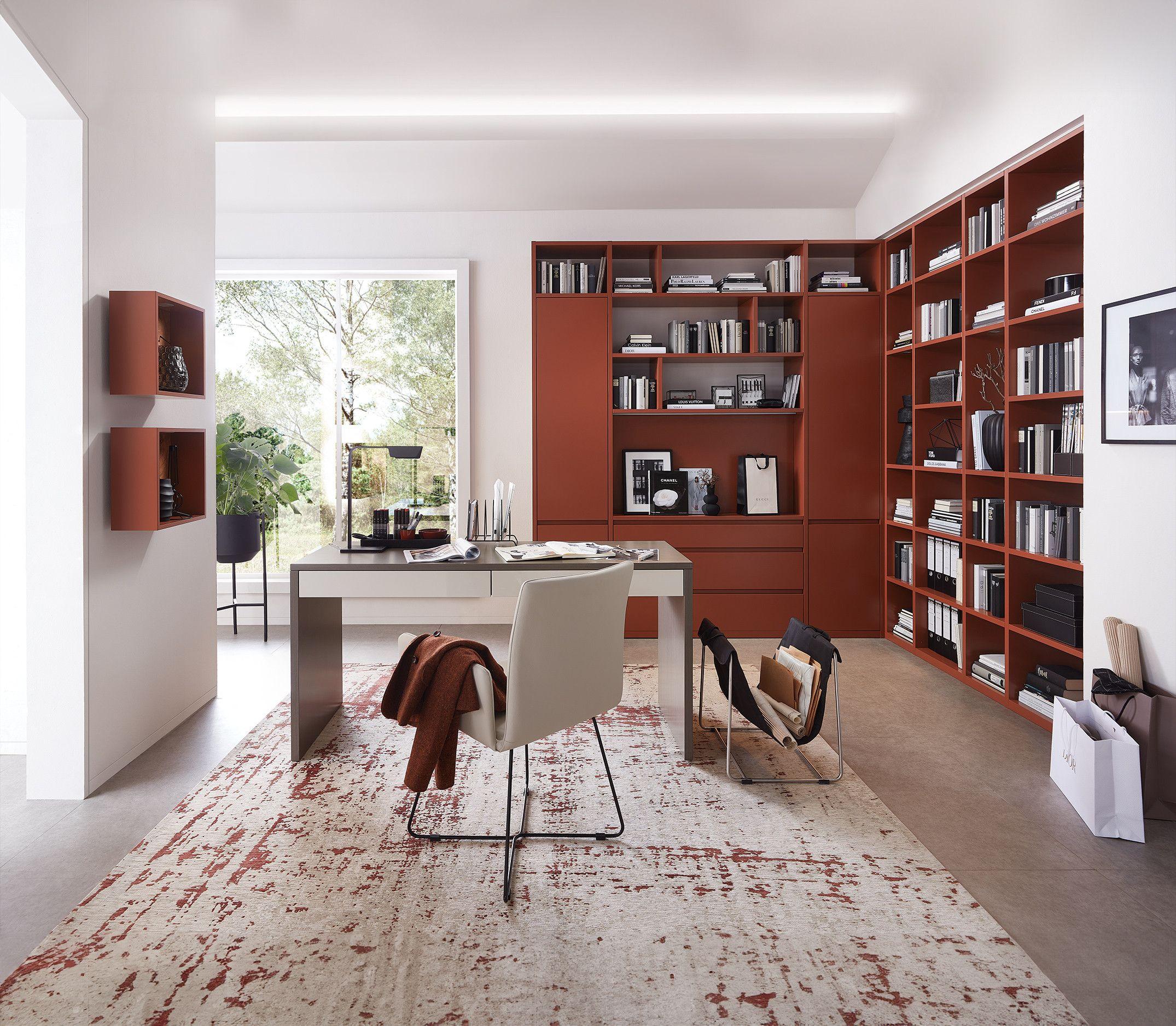 Helles, modernes Homeoffice mit warmroten Bücherregalen, einem zentralen Schreibtisch mit beiger Oberfläche und eleganten Möbeln. Große Fenster bieten Blick ins Grüne, während ein abstrakter Teppich in Rot- und Beigetönen den Raum akzentuiert.