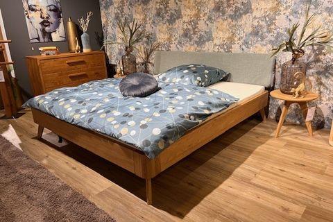 Doppelbett aus hellem Holz mit gepolstertem Kopfteil in Mintgrün, dekoriert mit blauer Bettwäsche mit Punktmuster. Links daneben eine Holzkommode, rechts ein runder Nachttisch mit Vase und goldener Affendekoration. Hintergrund mit floraler Tapete.