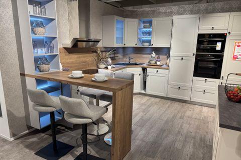 Weiße Landhausküche mit Rahmenfronten, Holzarbeitsplatte und Nischenrückwand in Eiche, Bartresen mit Polsterhockern, beleuchtetem Designregal mit blauer LED-Beleuchtung und Hochschrank mit Einbaugeräten.