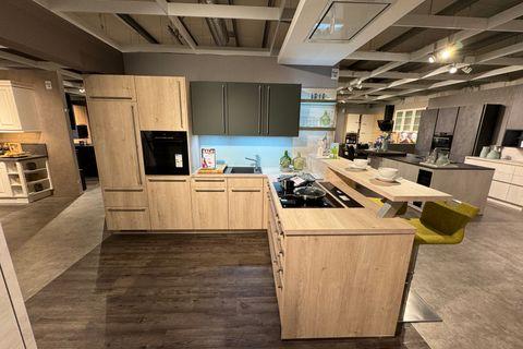 Küche in heller Eiche-Optik mit olivgrünen Oberschränken, Kochinsel mit integriertem Kochfeld und Bartresen, senfgelbem Barhocker, hellblauer Glasnischenrückwand und Hochschrank mit Einbaugeräten.