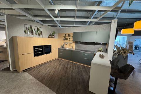 L-Küche in Metallic Ggrün und heller Eiche-Optik mit dunkler Arbeitsplatte, Holz-Nischenrückwand mit Relingsystem, Hochschrank mit Einbaugeräten, weißem Bartresen mit braunen Polsterhockern und dekorativer Blätter-Wanddeko.