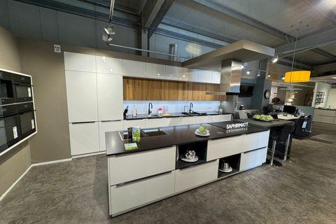 Große Küche in Saphirmatt Weiß mit dunkler Arbeitsplatte, Kochinsel mit Kochfeld und offenen Designfächern, Holz-Nischenrückwand in Eiche, grifflosen Fronten, Deckenlifthaube und integriertem Essplatz mit Barhockern.