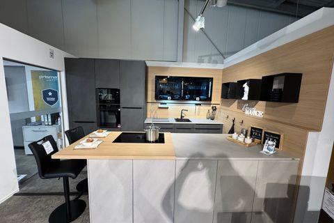 Moderne Küche in hellem Grau und Anthrazit mit Kochinsel, Holztresen und Barhockern, beleuchteter Glasvitrine, Eichen-Nischenrückwand mit schwarzen Wandregalen und Hochschrank mit Einbaugeräten.